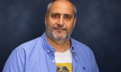 Πορτραίτο άνδρα με γκρίζα γένια, ανοιχτόχρωμο πουκάμισο και μπλουζάκι, κοιτάζει τον φακό με σοβαρό ύφος σε σκούρο φόντο.