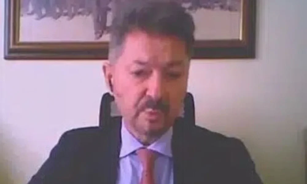 Άνδρας με κοστούμι και γραβάτα σε εσωτερικό χώρο με κάδρο στον τοίχο.
