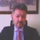 Άνδρας με κοστούμι και γραβάτα σε εσωτερικό χώρο με κάδρο στον τοίχο.