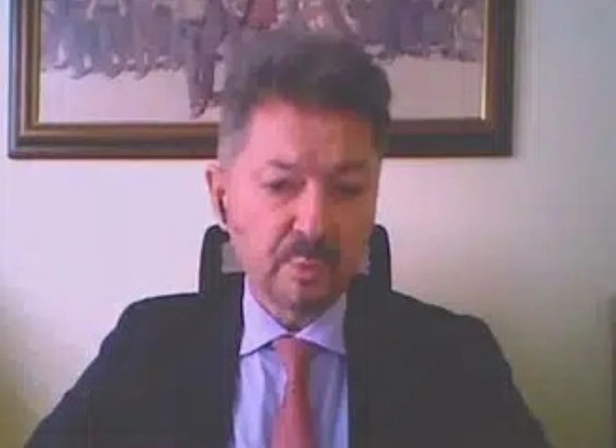 Άνδρας με κοστούμι και γραβάτα σε εσωτερικό χώρο με κάδρο στον τοίχο.