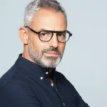 Άντρας με γυαλιά οράσεως, γκρίζα μαλλιά και μούσι φοράει σκούρο πουκάμισο σε ανοιχτόχρωμο φόντο