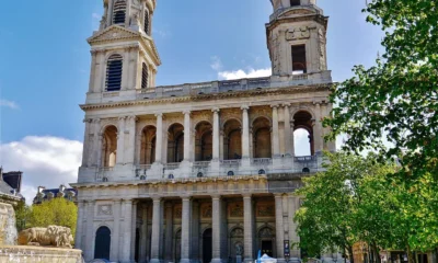 Πρόσοψη ιστορικού ναού με δύο πύργους και κιονοστοιχίες, μπροστά από πλατεία με συντριβάνι και δέντρα.