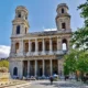 Πρόσοψη ιστορικού ναού με δύο πύργους και κιονοστοιχίες, μπροστά από πλατεία με συντριβάνι και δέντρα.