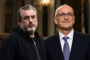 Δύο άνδρες στέκονται δίπλα-δίπλα, ο ένας φορά μαύρο φούτερ και ο άλλος σκούρο κοστούμι με γραβάτα.