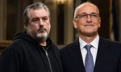 Δύο άνδρες στέκονται δίπλα-δίπλα, ο ένας φορά μαύρο φούτερ και ο άλλος σκούρο κοστούμι με γραβάτα.