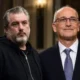 Δύο άνδρες στέκονται δίπλα-δίπλα, ο ένας φορά μαύρο φούτερ και ο άλλος σκούρο κοστούμι με γραβάτα.