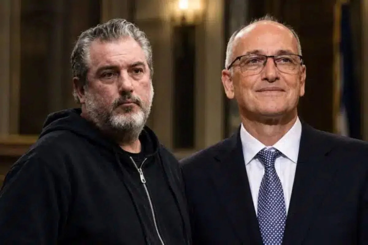 Δύο άνδρες στέκονται δίπλα-δίπλα, ο ένας φορά μαύρο φούτερ και ο άλλος σκούρο κοστούμι με γραβάτα.
