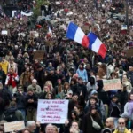 Πλήθος ανθρώπων να διαδηλώνει κρατώντας πλακάτ και γαλλικές σημαίες σε δρόμο.