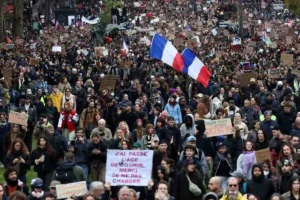 Πλήθος ανθρώπων να διαδηλώνει κρατώντας πλακάτ και γαλλικές σημαίες σε δρόμο.