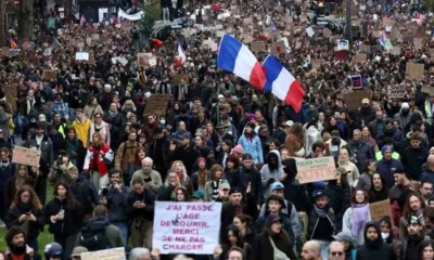 Πλήθος ανθρώπων να διαδηλώνει κρατώντας πλακάτ και γαλλικές σημαίες σε δρόμο.
