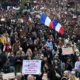 Πλήθος ανθρώπων να διαδηλώνει κρατώντας πλακάτ και γαλλικές σημαίες σε δρόμο.