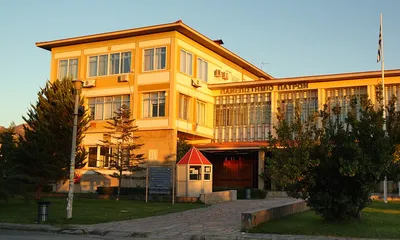 Εξωτερική άποψη κτιρίου με κίτρινη πρόσοψη, παράθυρα και ελληνική σημαία στον ιστό υπό το φως του ηλίου.