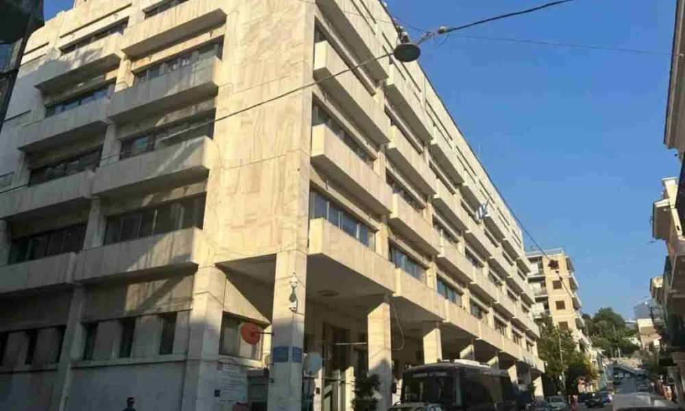 Πρόσοψη πολυώροφου κτιρίου σε δρόμο με σταθμευμένα αυτοκίνητα και πεζούς