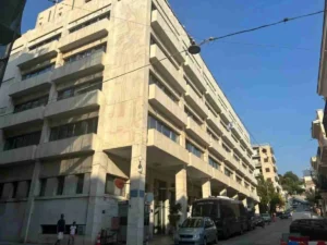 Πρόσοψη πολυώροφου κτιρίου σε δρόμο με σταθμευμένα αυτοκίνητα και πεζούς