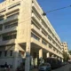 Πρόσοψη πολυώροφου κτιρίου σε δρόμο με σταθμευμένα αυτοκίνητα και πεζούς