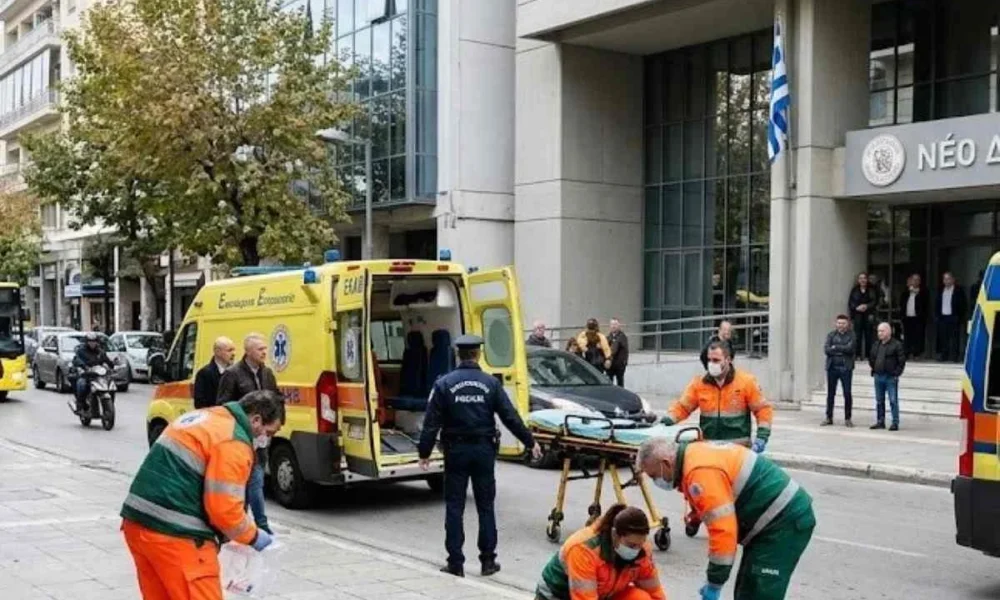 Διασώστες και αστυνομικός σε δρόμο μπροστά από κτήριο με ελληνική σημαία.