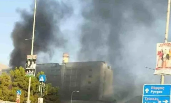 Πυκνοί μαύροι καπνοί υψώνονται πάνω από κτίριο κοντά σε δρόμο με αυτοκίνητα και πινακίδες.