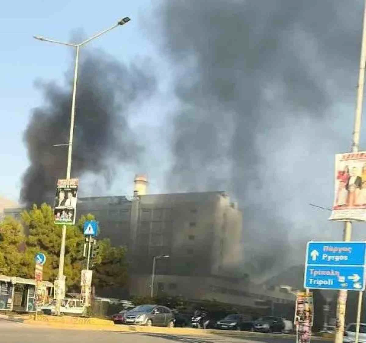 Πυκνοί μαύροι καπνοί υψώνονται πάνω από κτίριο κοντά σε δρόμο με αυτοκίνητα και πινακίδες.