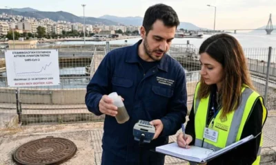 Δύο άτομα σε εξωτερικό χώρο με εγκαταστάσεις επεξεργασίας, κρατούν μπουκάλι με δείγμα και σημειωματάριο.