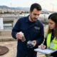Δύο άτομα σε εξωτερικό χώρο με εγκαταστάσεις επεξεργασίας, κρατούν μπουκάλι με δείγμα και σημειωματάριο.