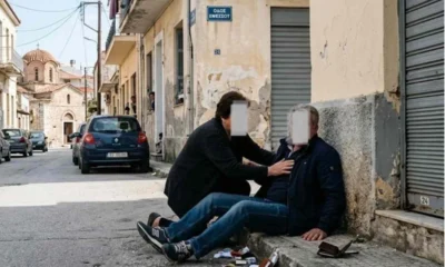 Άτομο καθισμένο στο πεζοδρομίο δίπλα σε ανοιχτά πορτοφόλια και αντικείμενα, ενώ άλλο άτομο σκύβει πάνω του σε δρόμο.