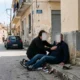 Άτομο καθισμένο στο πεζοδρομίο δίπλα σε ανοιχτά πορτοφόλια και αντικείμενα, ενώ άλλο άτομο σκύβει πάνω του σε δρόμο.