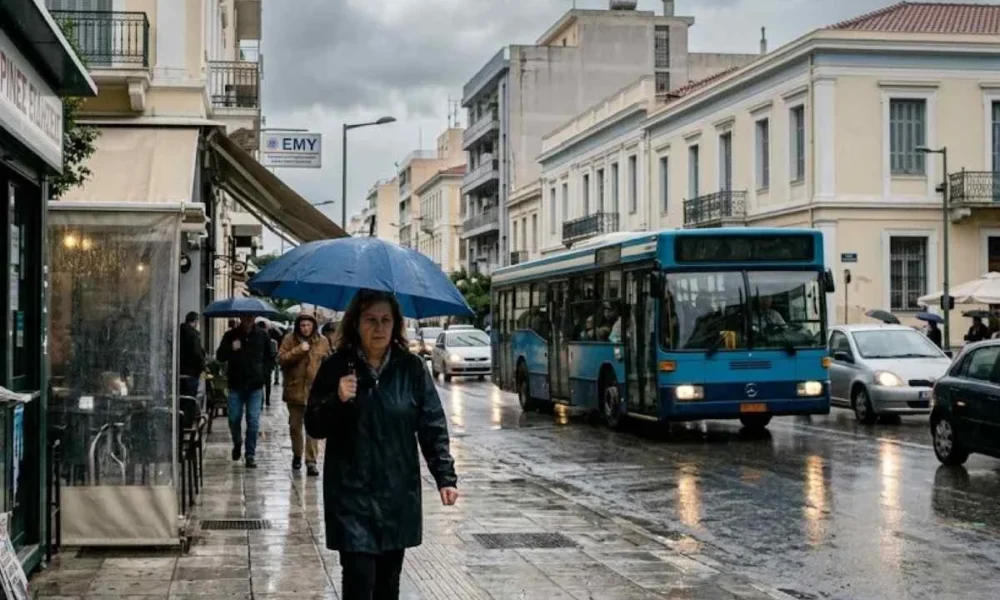Άνθρωποι με ομπρέλες περπατούν σε βρεγμένο δρόμο πόλης δίπλα σε λεωφορείο και αυτοκίνητα υπό συννεφιά