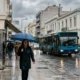 Άνθρωποι με ομπρέλες περπατούν σε βρεγμένο δρόμο πόλης δίπλα σε λεωφορείο και αυτοκίνητα υπό συννεφιά