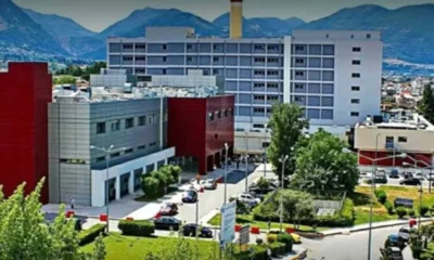 Εξωτερική άποψη πολυώροφου κτιρίου νοσοκομείου με πάρκινγκ, δέντρα και βουνά στο βάθος