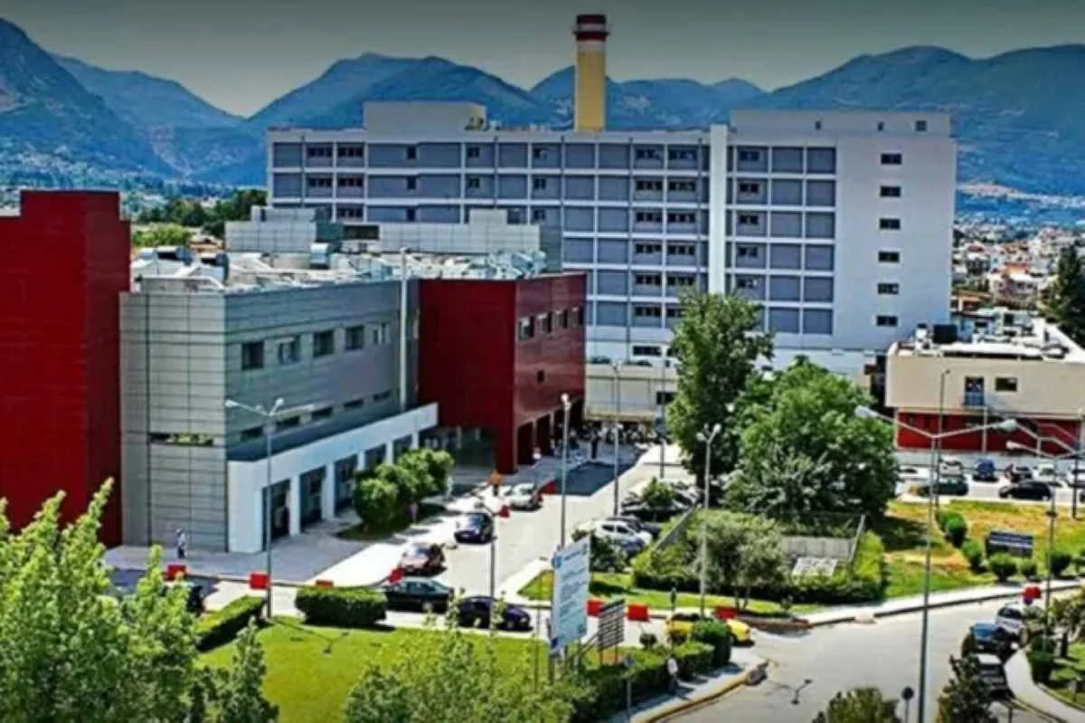 Εξωτερική άποψη πολυώροφου κτιρίου νοσοκομείου με πάρκινγκ, δέντρα και βουνά στο βάθος