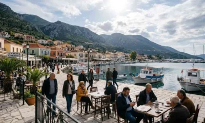 Άποψη παραλιακού οικισμού με σκάφη στο λιμάνι και άτομα σε τραπεζάκια καφετέριας.