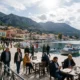 Άποψη παραλιακού οικισμού με σκάφη στο λιμάνι και άτομα σε τραπεζάκια καφετέριας.