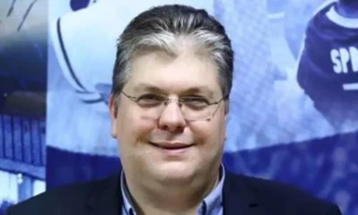 Άντρας με γυαλιά οράσεως και σκούρο σακάκι ποζάρει μπροστά σε μπλε φόντο.