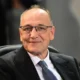 Άντρας με γυαλιά οράσεως, λευκό πουκάμισο, γραβάτα και σκούρο σακάκι καθισμένος σε κάθισμα.