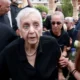 Γυναίκα με λευκά μαλλιά και σκούρα ρούχα περιστοιχισμένη από πλήθος σε εξωτερικό χώρο