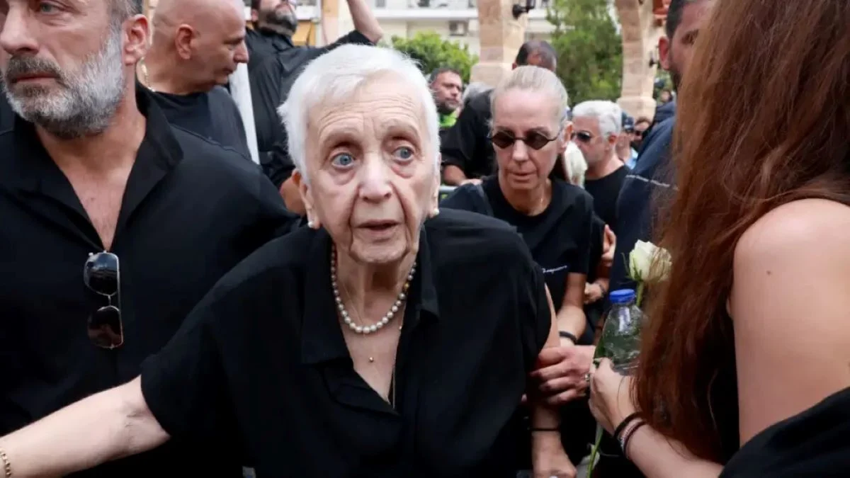 Γυναίκα με λευκά μαλλιά και σκούρα ρούχα περιστοιχισμένη από πλήθος σε εξωτερικό χώρο