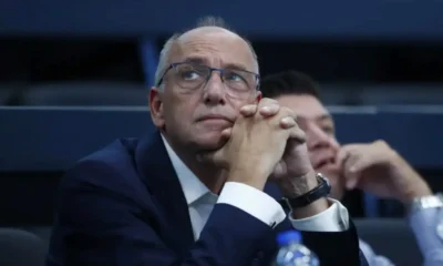 Άντρας με γυαλιά και σκούρο σακάκι ακουμπά το πηγούνι στα χέρια του σε εσωτερικό χώρο.