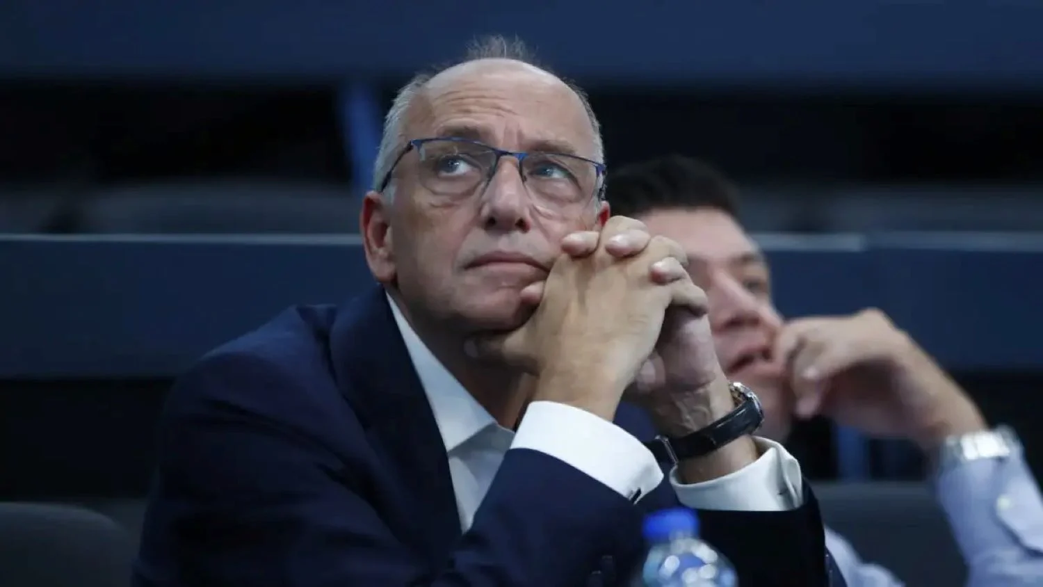 Άντρας με γυαλιά και σκούρο σακάκι ακουμπά το πηγούνι στα χέρια του σε εσωτερικό χώρο.