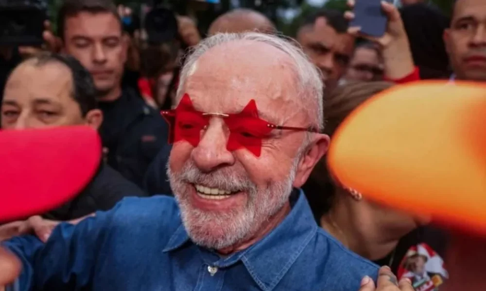 Ηλικιωμένος άνδρας με λευκά γένια και κόκκινα γυαλιά σε σχήμα αστεριού περιτριγυρισμένος από πλήθος.