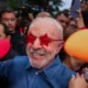Ηλικιωμένος άνδρας με λευκά γένια και κόκκινα γυαλιά σε σχήμα αστεριού περιτριγυρισμένος από πλήθος.