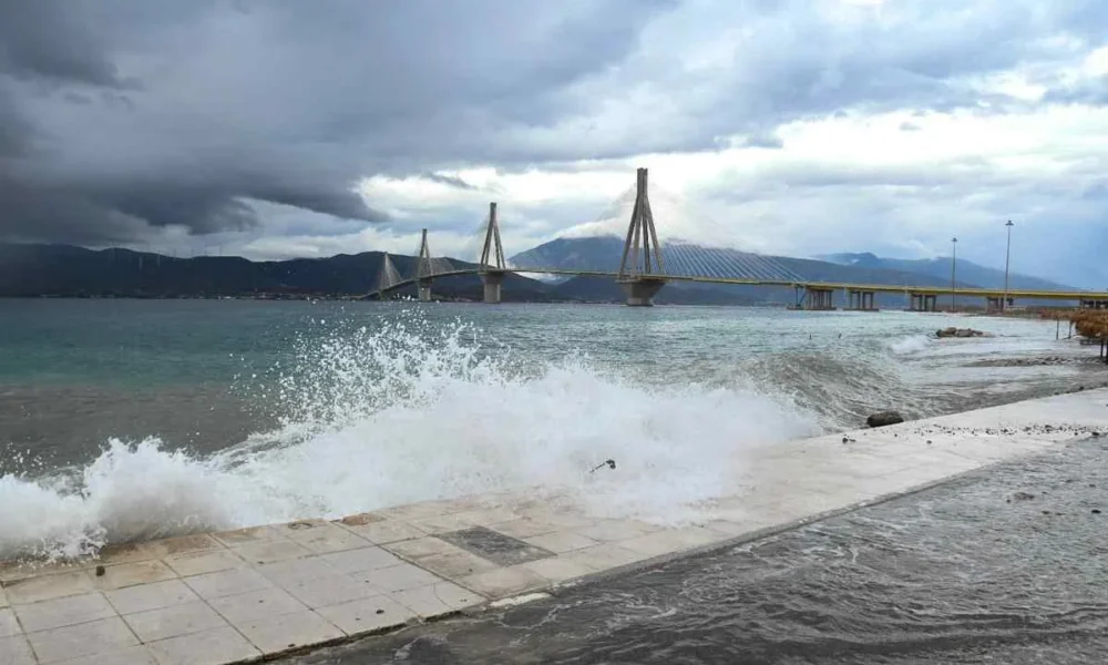 Θάλασσα με κύματα που σκάνε στην ακτή και γέφυρα στο βάθος κάτω από συννεφιασμένο ουρανό.