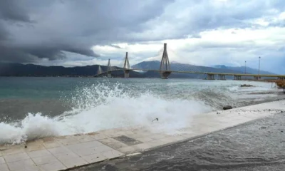 Θάλασσα με κύματα που σκάνε στην ακτή και γέφυρα στο βάθος κάτω από συννεφιασμένο ουρανό.