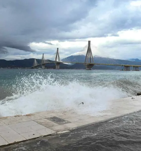 Θάλασσα με κύματα που σκάνε στην ακτή και γέφυρα στο βάθος κάτω από συννεφιασμένο ουρανό.