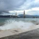Θάλασσα με κύματα που σκάνε στην ακτή και γέφυρα στο βάθος κάτω από συννεφιασμένο ουρανό.
