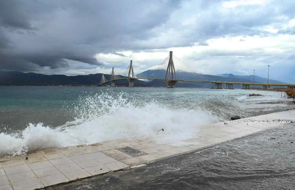 Θάλασσα με κύματα που σκάνε στην ακτή και γέφυρα στο βάθος κάτω από συννεφιασμένο ουρανό.