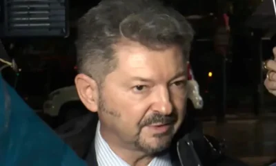 Άντρας με κοστούμι, γραβάτα και μούσι μιλάει περιστοιχισμένος από κόσμο.