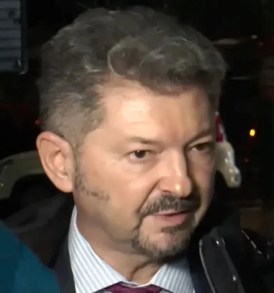 Άντρας με κοστούμι, γραβάτα και μούσι μιλάει περιστοιχισμένος από κόσμο.