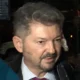 Άντρας με κοστούμι, γραβάτα και μούσι μιλάει περιστοιχισμένος από κόσμο.