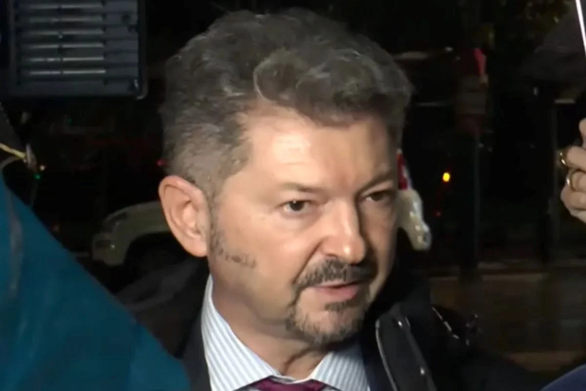 Άντρας με κοστούμι, γραβάτα και μούσι μιλάει περιστοιχισμένος από κόσμο.
