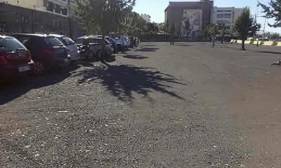 Ανοιχτός υπαίθριος χώρος στάθμευσης με παρκαρισμένα αυτοκίνητα στα αριστερά, δέντρα και κτίρια στο βάθος.
